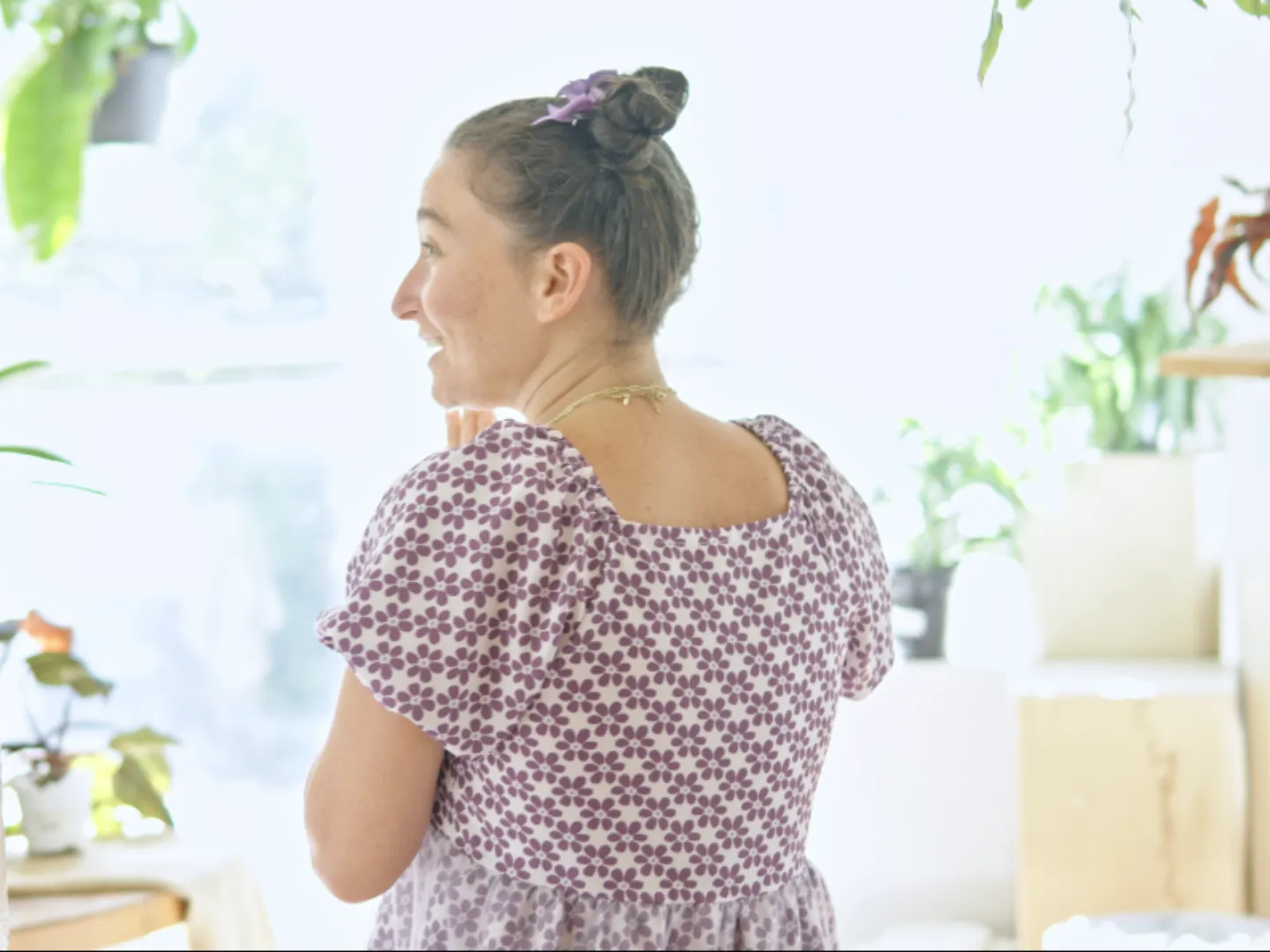 Woman in a purple floral dress laughing in a bright room filled with houseplants